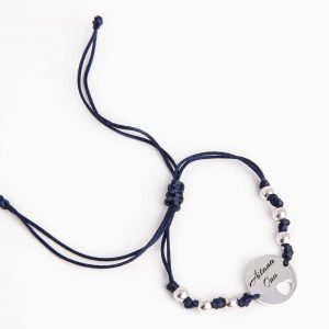 pulsera de cuerda con un trenzado de nudos y bolas de plata. Tiene una medalla redonda con un corazón calado y un grabado personalizado de dos nombres.